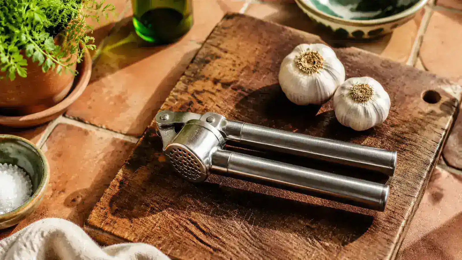 OXO Good Grips garlic press resting on a wood cutting board beside several whole garlic bulbs in warm kitchen light