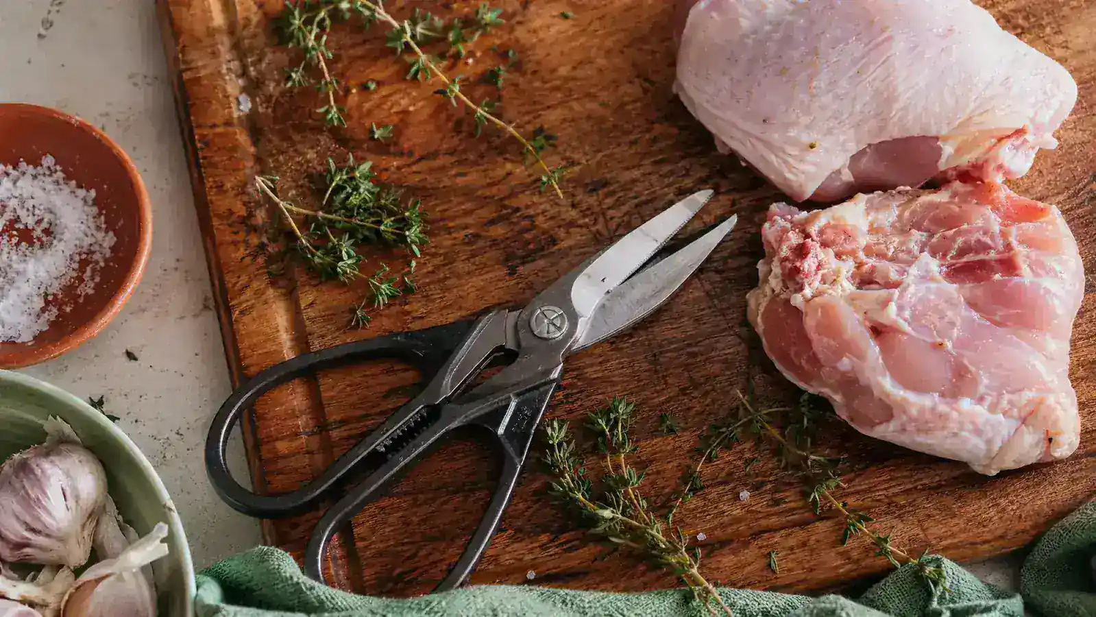 A pair of kitchen shears resting on a wooden cutting board next to fresh herbs and raw chicken