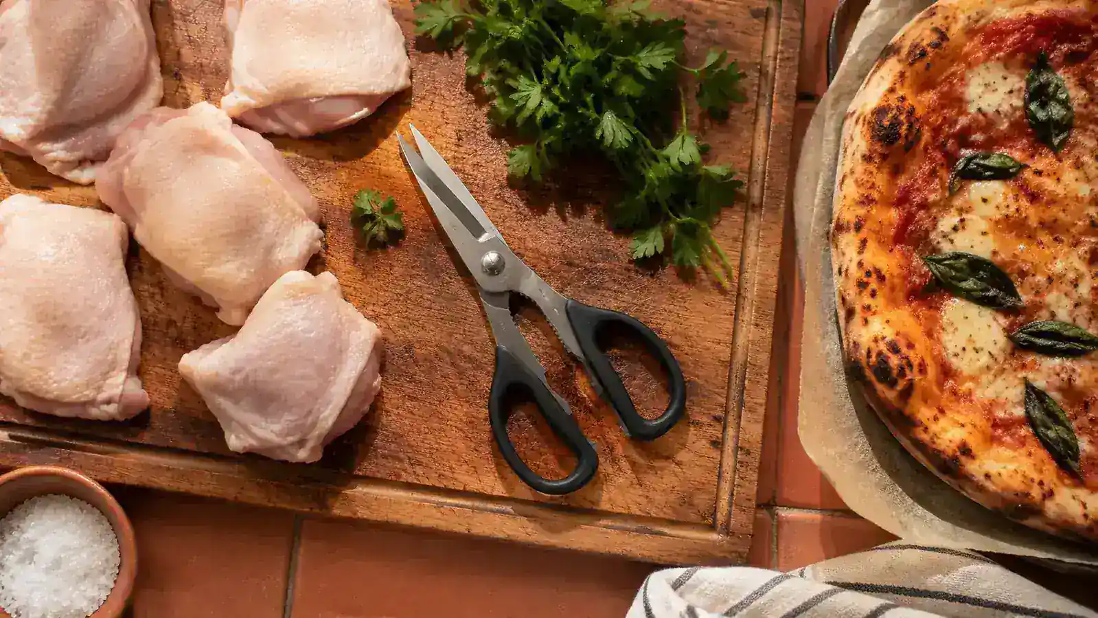 A pair of kitchen shears resting on a wooden cutting board next to raw chicken thighs, fresh herbs, and a pizza