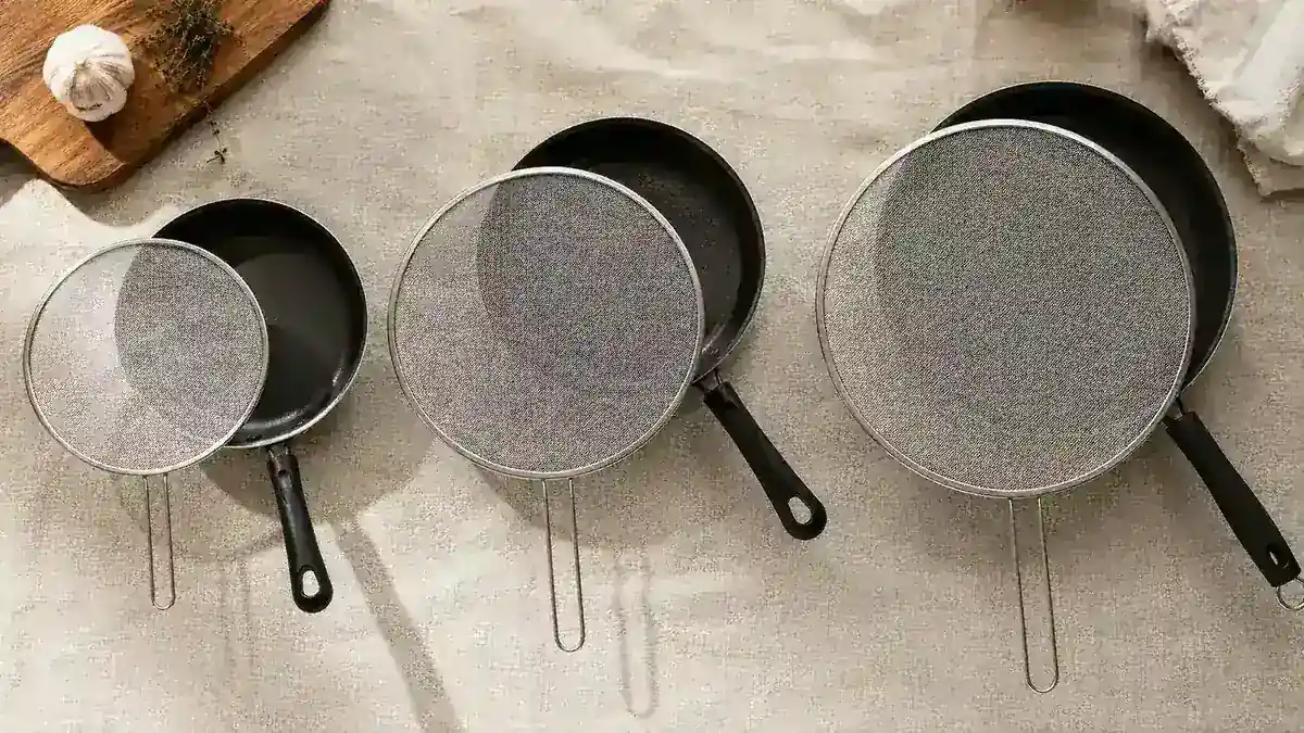 Three stainless steel splatter screens of different sizes laid out flat on a kitchen counter next to pans they match