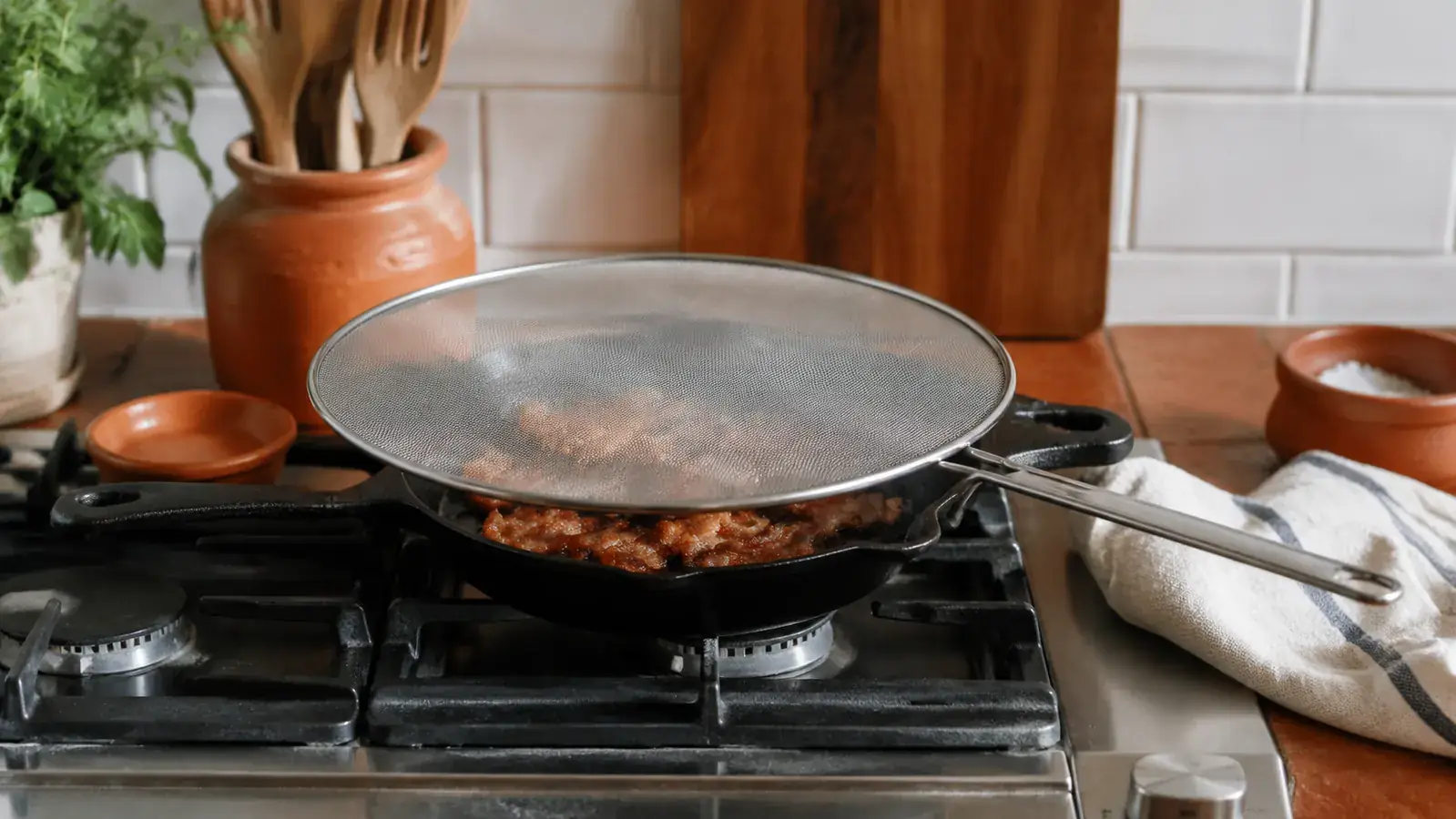 Cast iron skillet on a gas stovetop with a stainless steel splatter screen resting on top, bacon sizzling underneath, clean countertop visible in the background