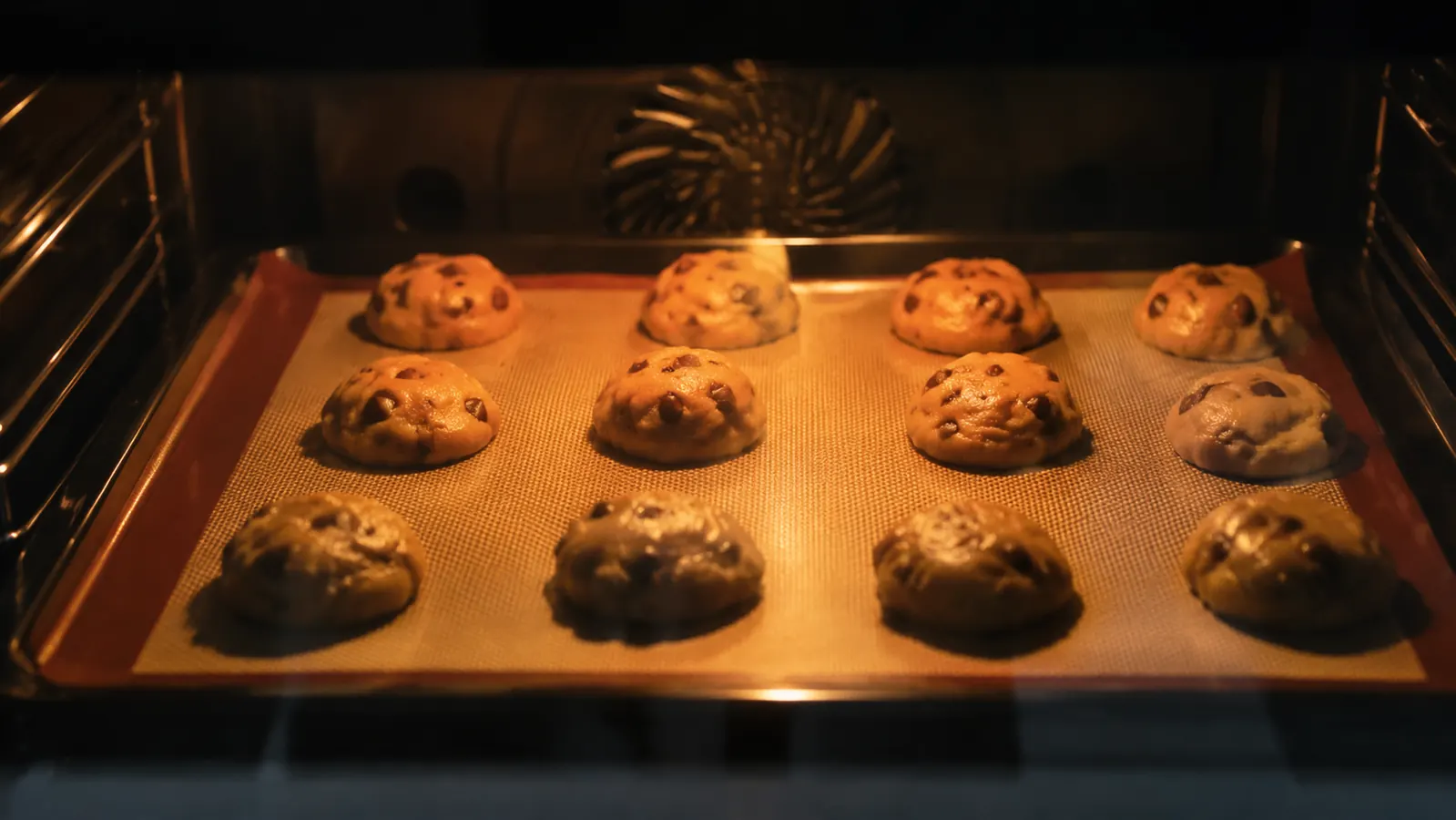 Cookie dough balls evenly spaced on a silicone mat inside an oven with oven light on
