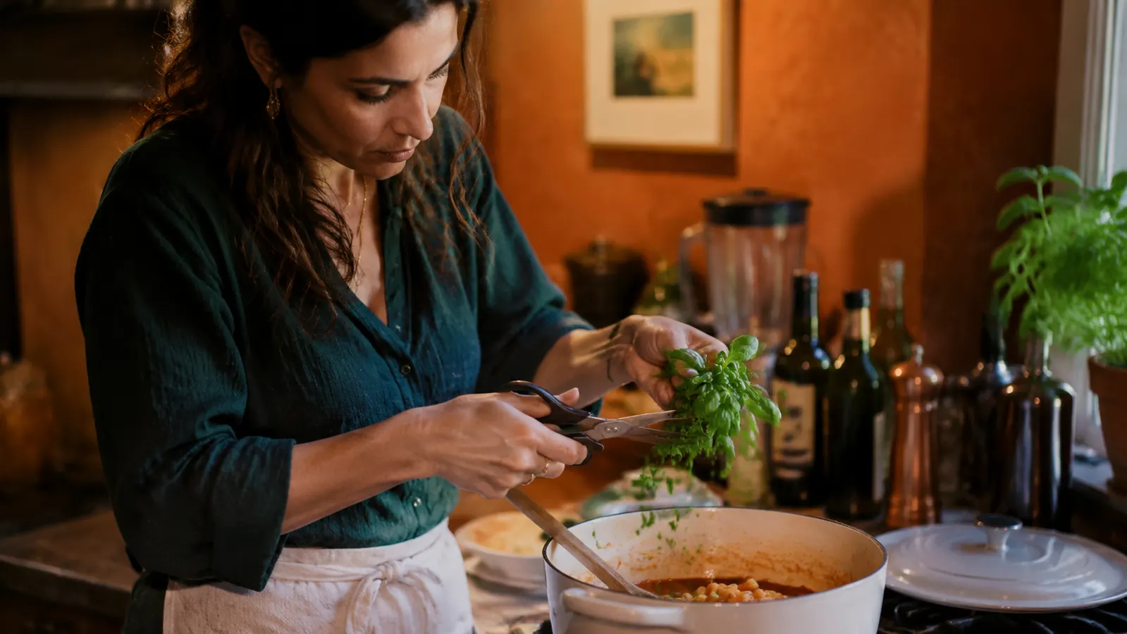 Woman snipping fresh herbs directly over a pasta pot on a gas stovetop, warm kitchen light
