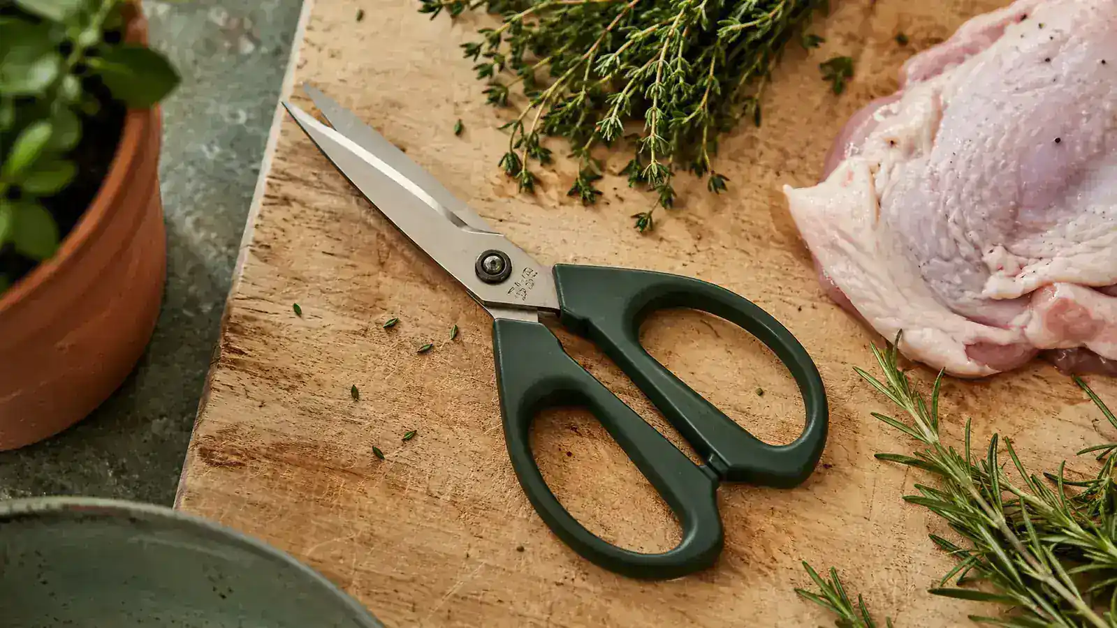 Pair of kitchen shears resting on a wooden cutting board beside fresh herbs and a raw chicken thigh, warm kitchen light