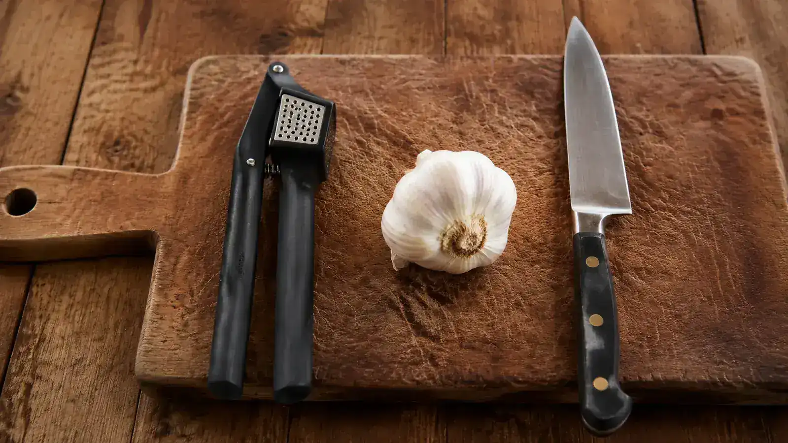 OXO garlic press resting on a wooden cutting board next to a head of garlic and a chef's knife