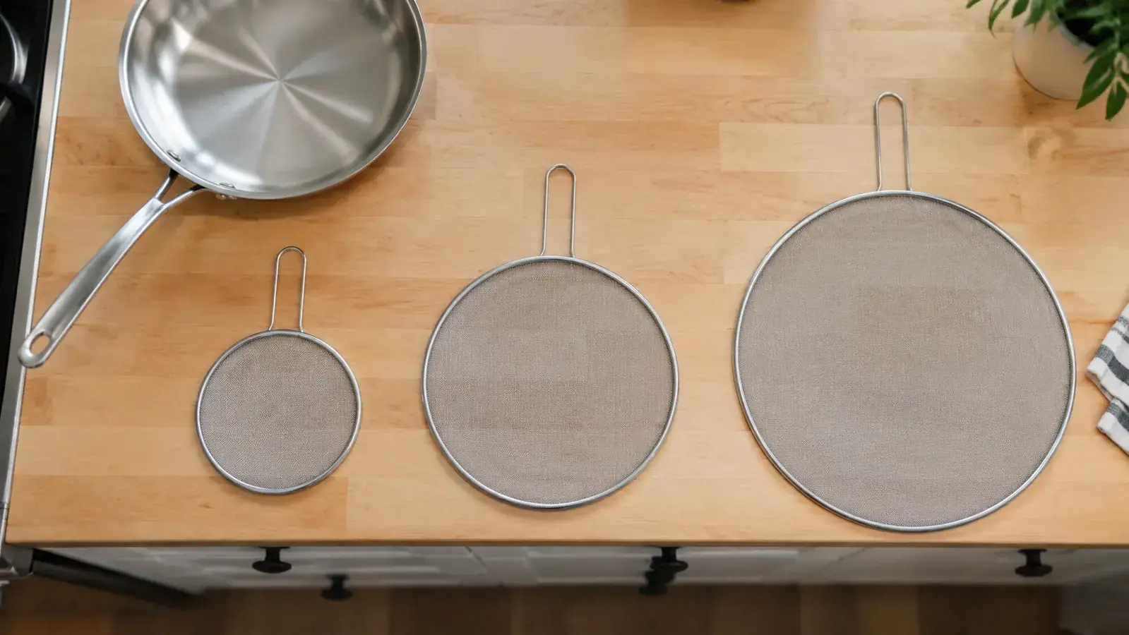 Three splatter screens of different sizes laid side by side on a clean wood kitchen counter next to a stainless pan